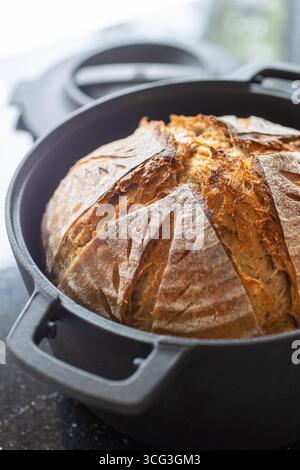 Pane a lievitazione naturale appena sfornato in un forno olandese in ghisa nera. La rustica crosta dorata presenta un motivo artistico che mette in risalto la lavorazione a mano Foto Stock