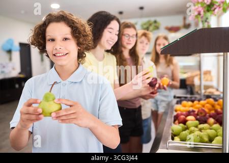 Gli studenti si riuniscono in una caffetteria scolastica pulita e ben organizzata, scegliendo tra le opzioni di frutta fresca da una vivace esposizione, promuovendo un'alimentazione sana tra le altre Foto Stock