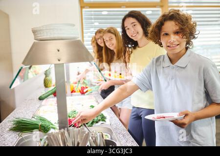 Un gruppo di studenti sorridenti viene servito cibo fresco e nutriente in una caffetteria scolastica. La scena cattura un'atmosfera amichevole con diversi indivi Foto Stock