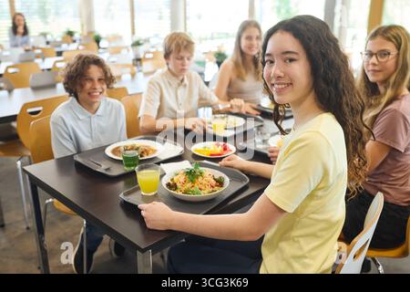 Gruppo di adolescenti seduti a un tavolo della caffetteria, gustando cibi e bevande salutari, mostrando cameratismo e un ambiente positivo in un ambiente pulito e pulito Foto Stock