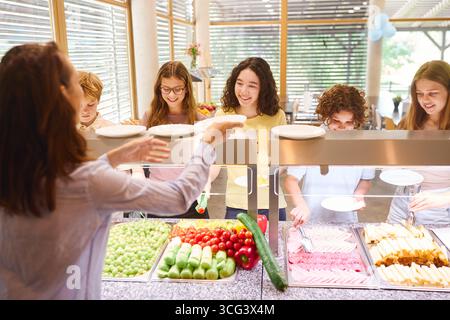 Gli studenti si riuniscono presso un bancone della caffetteria scolastica con una varietà di piatti freschi. Vengono serviti piatti e piatti selezionati. La sc Foto Stock