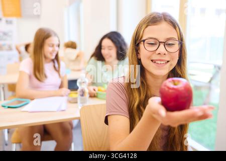 Un ambiente scolastico allegro con gli studenti che condividono un momento durante lo spuntino, concentrandosi su uno studente che ammira una mela rossa fresca in una classe ben illuminata Foto Stock