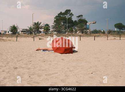 Una tranquilla spiaggia caratterizzata da un vibrante ombrello rosso che ombrega un sole su soffice sabbia dorata, circondato da lussureggianti palme e da un cielo nuvoloso. Foto Stock