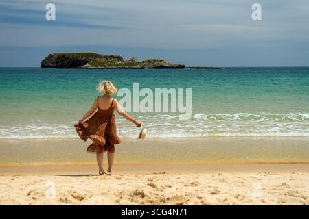 Sagres, Portogallo. 13 maggio 2017. Donna con un abito marrone che regge un cappello in piedi sulla spiaggia, guardando l'Oceano Atlantico turchese e la piccola isola in Foto Stock