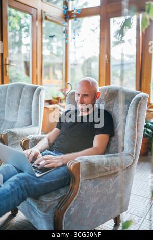 Uomo vestito con T-shirt nera e jeans seduto comodamente su una poltrona, che lavora su un computer portatile in casa, in salotto, in un soggiorno, in remoto Foto Stock