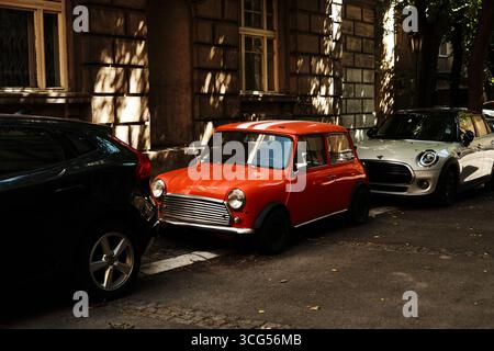 Auto Mini cooper rossa d'epoca parcheggiata tra veicoli moderni su una strada ombreggiata della città. Belgrado, Serbia - 26 agosto 2025. Foto Stock