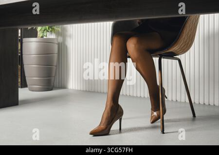 Gambe femminili sottili con collant abbronzato su una scrivania in un ufficio contemporaneo, con eleganti tacchi alti Foto Stock