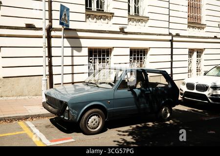 Vecchia auto blu Zastava Yugo parcheggiata su una strada soleggiata vicino a un edificio bianco. Belgrado, Serbia - 26 agosto 2025. Foto Stock
