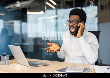 Un uomo d'affari seduto alla sua scrivania reagisce emotivamente durante una conversazione con il telefono cellulare. Il suo spazio di lavoro è dotato di un laptop, un telefono e un moderno ambiente di lavoro Foto Stock