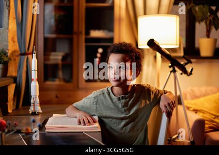 Ritratto di un ragazzo afroamericano felice seduto alla scrivania a casa a guardare lontano pensando ai pianeti di cui legge Foto Stock