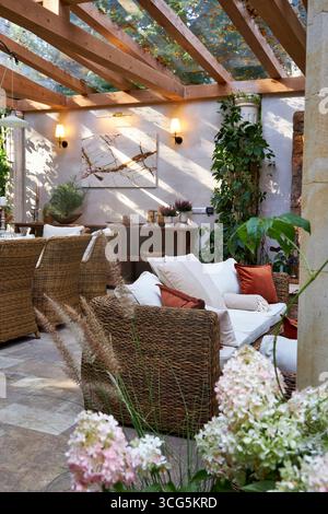 Elegante area salotto esterna con poltrone in vimini, cuscini bianchi e cuscini arancioni. Tavolo in legno decorato con candele, piante e dettagli autunnali Foto Stock
