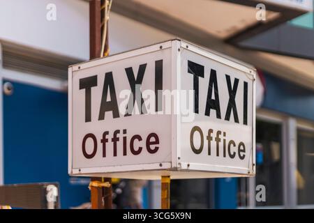 Un cartello dell'ufficio taxi si distingue con lettere in grassetto in un vivace ambiente urbano. Le persone sono probabilmente nelle vicinanze, impegnate in attività quotidiane e pendolari Foto Stock