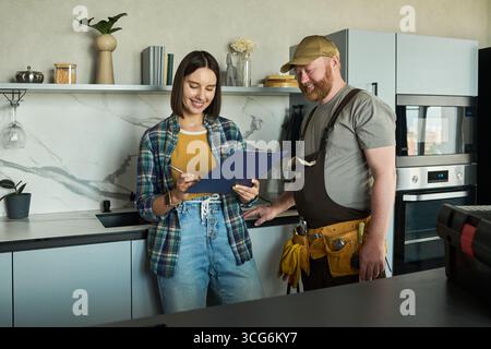 Giovane donna adulta caucasica che esamina documenti con uomo di mezza età caucasico che indossa uniformi da lavoro in cucina moderna, sia in piedi che sorridente mentre discute i dettagli del progetto Foto Stock