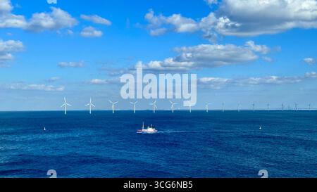 Parco eolico offshore con turbine all'orizzonte sotto il cielo blu, con barche in primo piano. Foto Stock