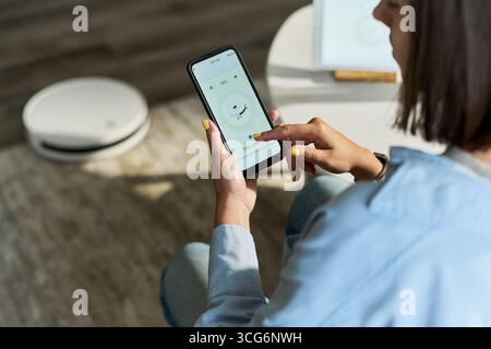 Giovane donna caucasica adulta che utilizza lo smartphone per controllare l'aspirapolvere robotizzato a casa, seduta a terra con interfaccia del dispositivo visibile sullo schermo, modalità Foto Stock