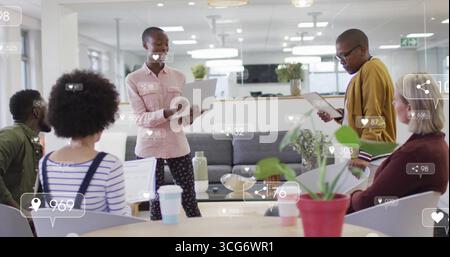 Donna che indossa una camicia rosa che regge un laptop nella moderna sala d'ufficio, con icone multimediali fluttuanti Foto Stock