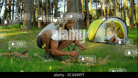Uomo accovacciato che accalca la legna da ardere nella fossa del fuoco nella bonifica della foresta con lo smartphone tenda zaino Foto Stock