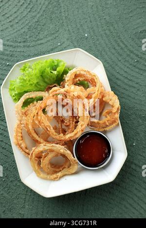 Abbondanza di anelli di cipolla fritti su piatto di ceramica, serviti con salsa piccante. Vista dall'alto, disposizione piatta Foto Stock