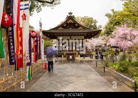 Un visitatore cammina lungo un sentiero in pietra verso un haiden tradizionale al Santuario di Hirano, il sentiero fiancheggiato da bandiere e striscioni colorate, Kyoto, Giappone Foto Stock