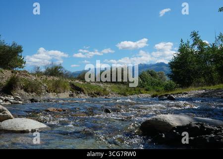 Veloce e vivace fiume con acqua fresca limpida che scorre su pietre con alberi sulle rive e montagne e cielo blu in lontananza nelle soleggiate giornate estive in TR Foto Stock