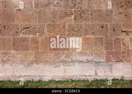 Vecchio muro in pietra che mostra la sua consistenza resistente agli agenti atmosferici e le tonalità naturali della terra, mattoni di tufo rosso e erba verde, sfondo interno astratto Foto Stock