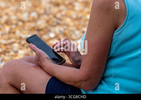 primo piano di una donna anziana che utilizza uno smartphone o un telefono cellulare con abbigliamento estivo. donna anziana seduta sulla spiaggia utilizzando uno smartphone o un telefono cellulare Foto Stock