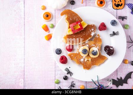 Sorpresa gastronomica per bambini di Halloween. Divertenti pancake in forma di simpatico pipistrello con cappello da strega. Spuntino a colazione per la festa di Halloween dei bambini, con frutti di bosco e caramelle Foto Stock