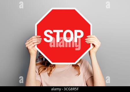 Donna che tiene in mano un poster con un cartello di stop che copre il viso isolato su sfondo grigio Foto Stock