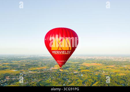 Sint-Martens-Latem, Belgio - 25 agosto 2025: Vista aerea di una vivace mongolfiera rossa impreziosita da "Mervielde" che galleggia dolcemente sopra il lussureggiante paesaggio verde. Foto Stock