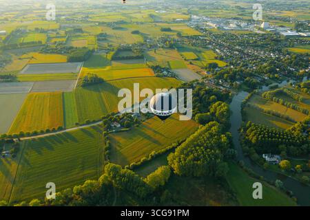 Sint-Martens-Latem, Belgio - 25 agosto 2025: Vista aerea di una mongolfiera che si sposta su campi verdeggianti e il tortuoso fiume Leie, un arazzo di verdi e gialli sotto la luce soffusa del mattino. Foto Stock