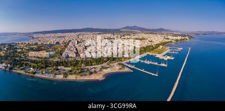 Vista aerea del lungomare orientale di Salonicco con porticciolo, edifici urbani, vegetazione lussureggiante e montagne lontane sotto un cielo limpido. Foto Stock