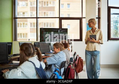 Donna caucasica di mezza età che tiene un tablet in piedi vicino a un gruppo di bambini diversi seduti alla scrivania utilizzando computer desktop in aula, insegnante che osserva gli studenti che lavorano Foto Stock