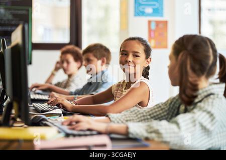 Gruppo di bambini caucasici e multietnici preadolescenti seduti alla scrivania utilizzando computer desktop in classe, ragazza in primo piano che gira e sorride al compagno di classe mentre digita sulla tastiera Foto Stock