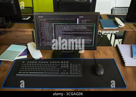 Monitor per computer che visualizza l'editor di codice con script di programmazione sullo schermo, tastiera e mouse standard posizionati sulla scrivania in aula, notebook e cancelleria visibili nelle vicinanze Foto Stock