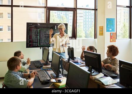 Donna caucasica di mezza età che insegna un gruppo di bambini diversi in classe di computer, in piedi vicino a un display interattivo, tenendo un tablet, studenti che alzano le mani, monitor sulle scrivanie Foto Stock