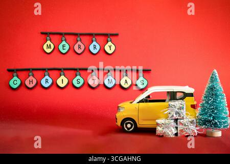 Scena natalizia con una piccola macchinetta giocattolo gialla, regali di Natale avvolti in argento, un mini albero verde e le parole "buon Natale" in lettere colorate Foto Stock