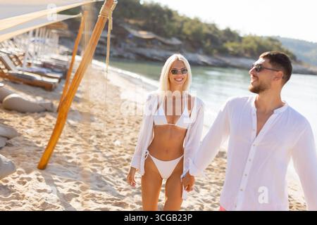 Una coppia felice passeggia lungo una spiaggia sabbiosa sotto il sole, abbracciando il momento in un elegante abbigliamento da spiaggia mentre si godono la compagnia reciproca. Foto Stock