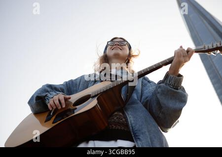 Giovane donna caucasica che suona la chitarra acustica all'aperto sorridendo verso l'alto Foto Stock