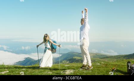 L'uomo festeggia con le braccia sollevate mentre la donna si erge con pali da trekking sulla cima erbosa della montagna sopra le nuvole. Foto Stock