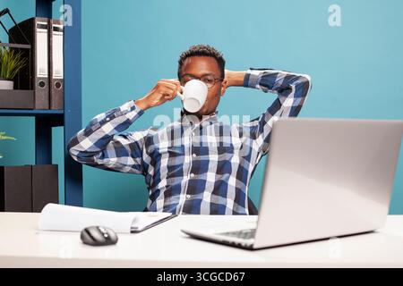 Uomo afroamericano che si rilassa sulla sedia e beve un caffè mentre guarda un video sul suo portatile. Giovane seduto alla scrivania, appoggiato all'indietro con una tazza in mano e guardando lo schermo del dispositivo. Foto Stock