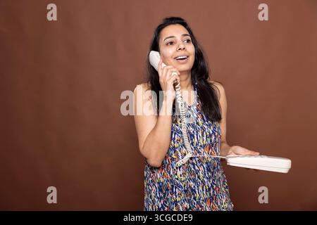 Una donna indiana sorridente tiene il ricevitore del telefono all'orecchio, parlando felicemente con i propri cari in studio. Joyful female individual utilizza la linea fissa per le telefonate, emettendo un sorriso brillante. Foto Stock