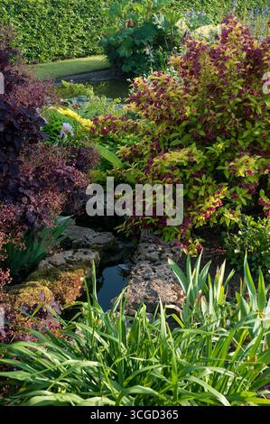 Un lussureggiante giardino presenta una gamma di piante dai colori vivaci che circondano un piccolo laghetto riflettente, creando un'atmosfera tranquilla in primavera. Foto Stock