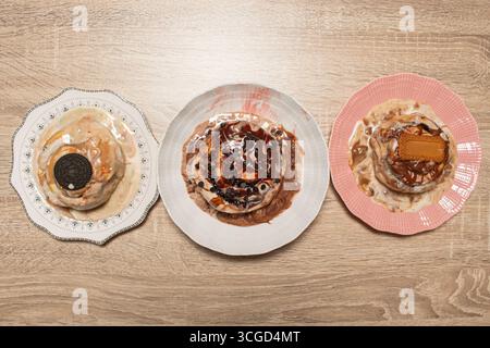 Vista dall'alto verso il basso di un assortimento di tre panini alla cannella gourmet su piatti colorati con diverse guarnizioni dolci su un solido sfondo di legno chiaro Foto Stock