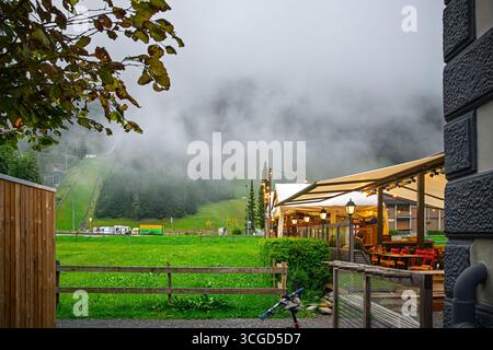 Una caffetteria illuminata sulle colline ai piedi del monte Titlis nella città di Engelberg, Svizzera, Alpi svizzere, con una funicolare di montagna alle spalle mentre la nebbia scorre Foto Stock
