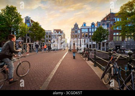 Biciclette e pedoni su un ponte ad anello interno del canale nel centro storico di Amsterdam in autunno, mentre il sole inizia a tramontare. Foto Stock