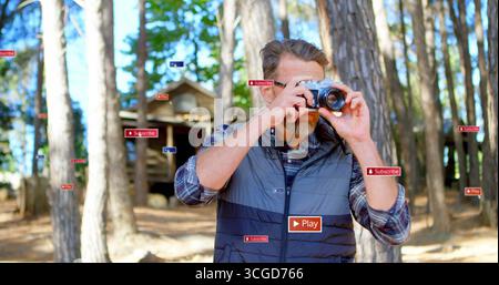 Inquadra un fotografo maturo in gilet blu che regola la fotocamera mirrorless nella pineta, con cabina Foto Stock