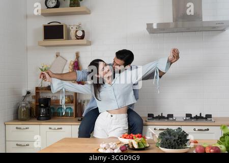 Giovane coppia romantica ballando gioiosamente in cucina moderna, simboleggiando l'amore, legame familiare, benessere, stile di vita sano, cibo biologico, insieme, h Foto Stock