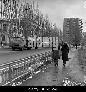 Una fotografia archivistica in bianco e nero della vita cittadina di Sloviansk all'inizio degli anni '1980, che cattura Svobody Street durante la bassa stagione con un flusso di camion e pedoni sovietici GAZ sullo sfondo di un nuovo edificio di nove piani, che riflette i contrasti della pianificazione urbana sovietica e la vita quotidiana dell'era della stagnazione, che serve come un prezioso documento della vita pacifica nel Donbas prima della guerra Foto Stock