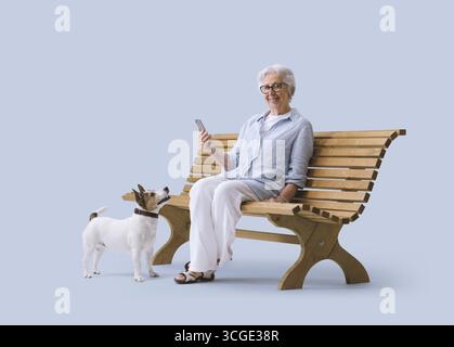 Donna anziana sorridente e felice con il telefono seduto su una panchina con il suo cane che guarda la macchina fotografica Foto Stock