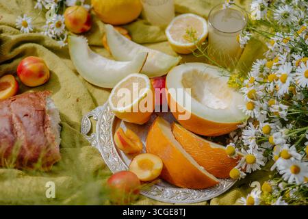 Un vivace spettacolo di meloni freschi, pesche e limoni è collocato su un piatto d'argento tra fiori colorati ed elementi naturali, creando un allegro bancomat Foto Stock